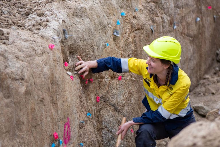 A woman in an Australian Government high-viz shirt and hard hat inspects marked areas of a rock wall.