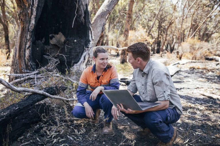 Woman in orange top squats next to man with laptop in Australian bush landscape