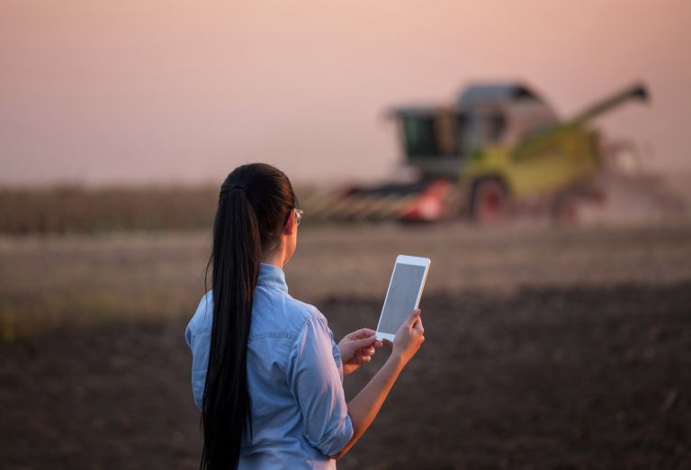 Woman with long dark hair holds computer tablet up to farm tractor in distance 