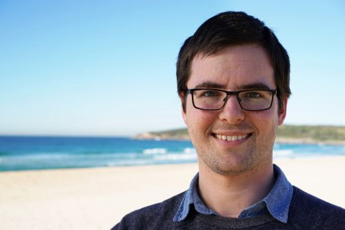 Smiling man with glasses stands in front of beach and looks to camera